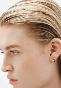 Pendiente de aro dorado con un pequeño acento de gema roja, diseño circular, textura suave, llevado en un tono de piel neutro.