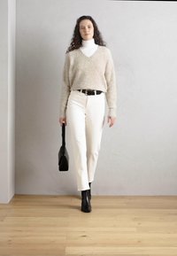 Beige, teksturert genser med V-hals, parret med hvite smale jeans og svarte ankellester. En liten svart veske holdes i høyre hånd.