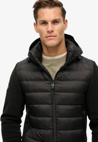 Sort puffer vest med lynlås, kombineret med et mørkt fleece-materiale. Har en hætte og logo-patch på venstre ærme.