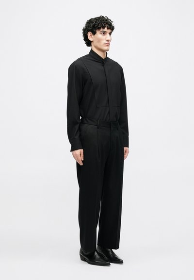 Μαύρο μακρυμάνικο πουκάμισο με υψηλό γιακά και πλισέ σχέδιο, συνδυασμένο με χαλαρά μαύρα παντελόνια και μυτερά μαύρα παπούτσια.