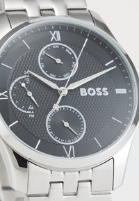 Montre en acier inoxydable argenté avec un cadran noir texturé, des aiguilles argentées, des sous-cadrans pour la date et le jour de la semaine, arborant un logo "BOSS" bien en vue.