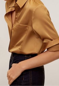 Chemise en satin à manches courtes de couleur jaune doré avec un col et une poche poitrine simple, portée avec un jean en denim foncé. Texture lisse visible.