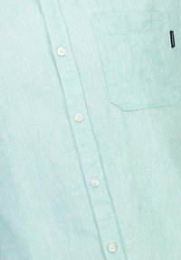 Camicia button-up verde chiaro realizzata in tessuto texturizzato, con taschino sul petto sinistro e cinque bottoni bianchi lungo la parte anteriore.