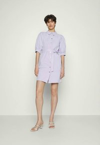 Vestido camisa de cor lilás claro com mangas curtas e bufantes, frente com botões e cintura com laço. Tecido suave, design até ao joelho e acessórios simples.