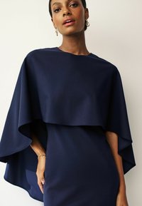 Mörkblå klänning med en lager-på-lager kapdesign, tillverkad av mjukt material. Har en rundad halsringning och subtilt drapering, med ett armband på handleden.