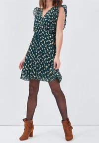 Robe fleurie verte avec manches à volants et col en V, dotée d'une jupe plissée. Portée avec des bottines en daim marron et des collants noirs.