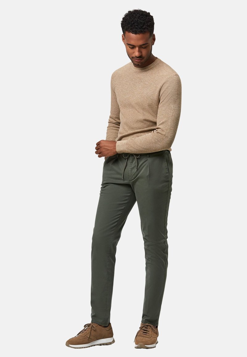 Pull beige texturé associé à un pantalon olive vert cintré, avec une taille élastique, et des baskets brunes avec des accents blancs.