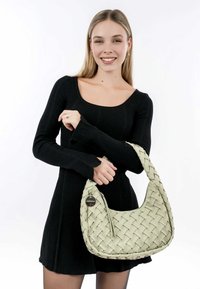Geweven groene handtas met een gebogen vorm, voorzien van een ritssluiting, textuurpatroon en een decoratieve tag. Gestyled met een zwarte jurk.