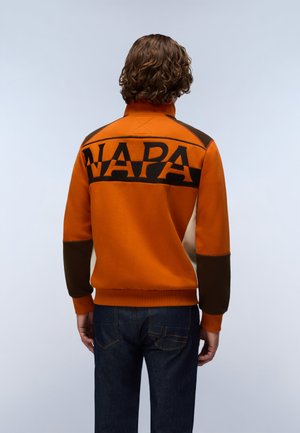 Persoon met krullend haar die een oranje en bruine jas draagt met "NAPA" gedrukt over de bovenrug, gecombineerd met donkerblauwe spijkerbroek.