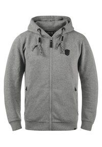 Siva zip-up dukserica napravljena od mekanog materijala, sa kapuljačom, dva bočna džepa i crnim detaljima. Uključuje pertle s metalnim vrhovima.