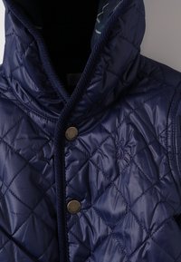 Gilet trapuntato blu navy con colletto alto, chiusura a scatto e logo ricamato. Presenta un'esterno liscio e lucido e un fodero testurizzato in contrasto.