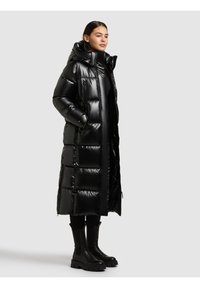 Melnā pūkaina jaka ar kapuci, ar garu quiltētu dizainu un spīdīgu virsmu. Valkāta kopā ar melnām potīšu zābakām un legingiem.