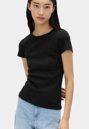 Ung kvinde med langt sort hår iført en tætsiddende sort t-shirt med korte ærmer og lyseblå jeans, stående foran en ensfarvet baggrund.