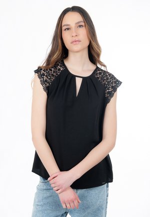 Jonge vrouw met lang haar, gekleed in een zwart topje met kanten mouwen en lichtblauwe spijkerbroek, staand met samengevouwen handen voor zich op een witte achtergrond.