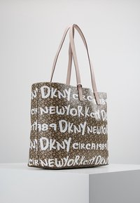 Sac fourre-tout marron avec des poignées rose clair, couvert de texte blanc en style graffiti lisant "DKNY", "New York" et "Circa 1989", posé sur un socle blanc.