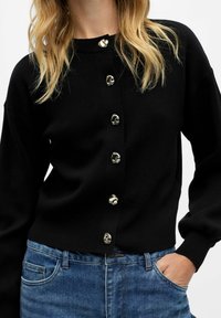 Cardigan nero in maglia con collo rotondo, dotato di sei bottoni dorati lucidi sul davanti. Abbinato a jeans in denim blu, dalla vestibilità casual.