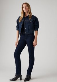 Chaqueta de mezclilla en azul oscuro con un diseño cropped, cierre de botones y bolsillos clásicos. Jeans oscuros ajustados, combinados con botas de tobillo negras con tacón.