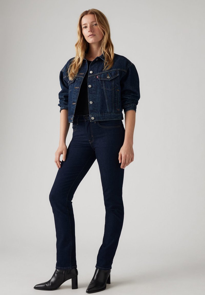 Chaqueta de mezclilla en azul oscuro con un diseño cropped, cierre de botones y bolsillos clásicos. Jeans oscuros ajustados, combinados con botas de tobillo negras con tacón.