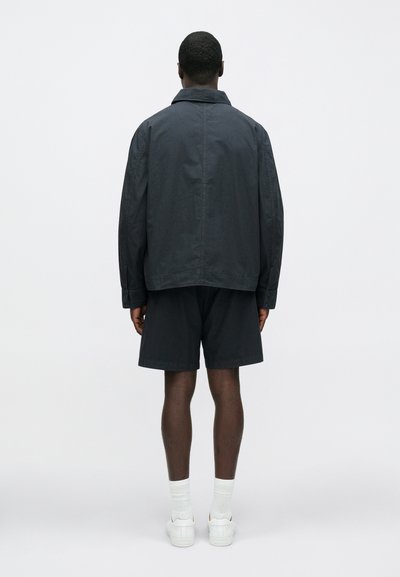 Ciemna, lekka kurtka wierzchnia z kołnierzem dołożonym na płasko, w zestawieniu z ciemnymi szortami. Gładka faktura, minimalistyczny design i luźny krój.