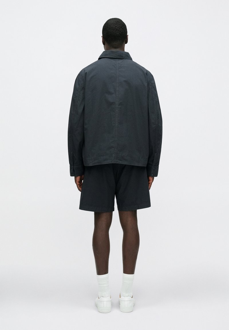 Dark, lightweight outerwear jacket with a fold-down collar, paired with dark shorts. Smooth texture, minimal design, and relaxed fit.