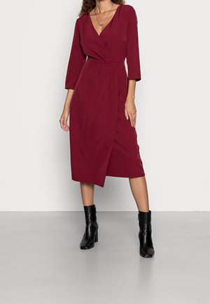 Femme portant une robe portefeuille bordeaux mi-longue avec des manches trois-quarts et des bottines noires, debout sur un sol gris devant un fond blanc.
