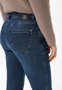 Donkerblauwe denim jeans hebben een aansluitend ontwerp met achterzakken, een gestructureerde tailleband en een leren label op de achterkant. Glad oppervlak met stikseldetails.