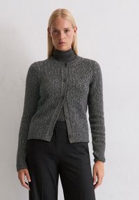 Cardigan grigio lavorato a maglia con tessuto testurizzato, scollatura rotonda e chiusura con bottoni. Indossato sopra un dolcevita scuro e abbinato a pantaloni a righe.