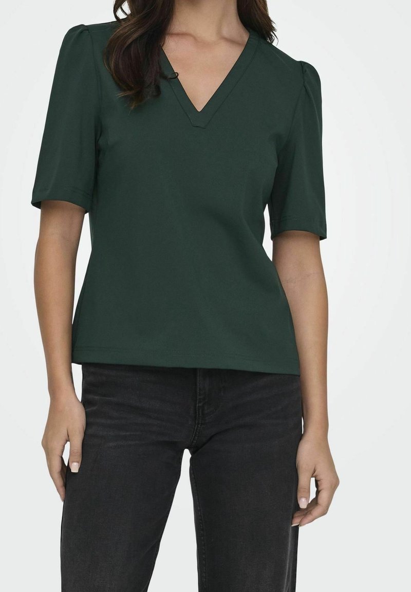 Femme portant un haut vert foncé à manches courtes et col en V ainsi qu'un jean noir, debout devant un fond clair uni.
