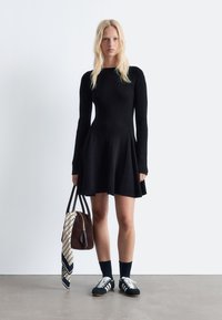 Schwarzes geripptes Kleid mit langen Ärmeln und ausgestellt Rock, kombiniert mit einer braunen Tasche und einem gemusterten Schal. Schwarze Socken und Sneakers runden den Look ab.
