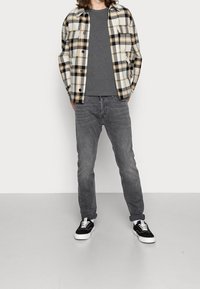 Person trägt ein schwarz-weiß kariertes Hemd über einem grauen T-Shirt, graue Jeans und schwarze Sneakers mit weißen Sohlen vor einfarbigen Hintergrund.