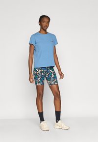 Μπλε κοντομάνικη κοντή μπλούζα συνδυασμένη με ποδηλατικό σορτς με σχέδια φύλλων σε πράσινο, λευκό και μπλε. Αθλητικά παπούτσια σε ουδέτερους τόνους.