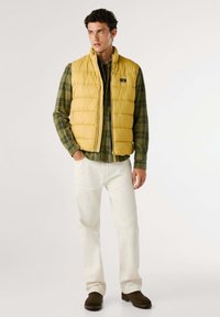 Gilet imbottito giallo sopra una camicia a quadri verde, abbinato a pantaloni bianchi e stivali marroni. Texture liscia con cuciture visibili e tasche.