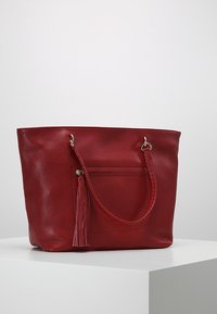 Czerwona skórzana torba tote o teksturowanym wykończeniu, płaskim dnie, długich uchwytach i akcentem w postaci frędzla, z przednią kieszenią na zamek.