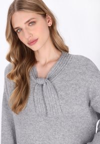 Pulover gri tricotat, cu o guler ribat și detaliu nod. Textură moale și croială relaxată, cu mâneci lungi și fără accesorii vizibile.