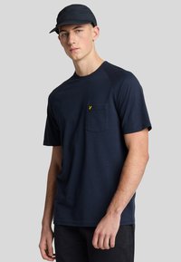 Maglietta blu navy con maniche corte, scollatura rotonda e taschino sul petto con un piccolo logo giallo. Tessuto morbido, vestibilità comoda.