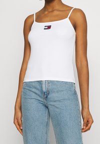 Vit bomullstanktop med tunna band som har en liten svart, rosa och vit rektangulär logotyp. Bärs med ljusblå denimjeans.