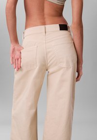 Beige wijde broek met hoge taille, voorzien van twee achterzakken en subtiele stikdetails. Glad katoenen stof.