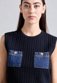 Ung kvinne med langt, mørkt hår har på seg en ermeløs, rippet svart topp med to denim-brystlommer festet med gullknapper.