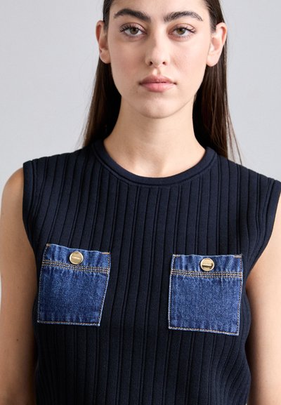 Noor naine pikkade tumedate juustega kannab musta varrukateta ribakanga topi, millel on kaks denimpolo taskut, mille kinnituseks on kuldsetest nuppudest.
