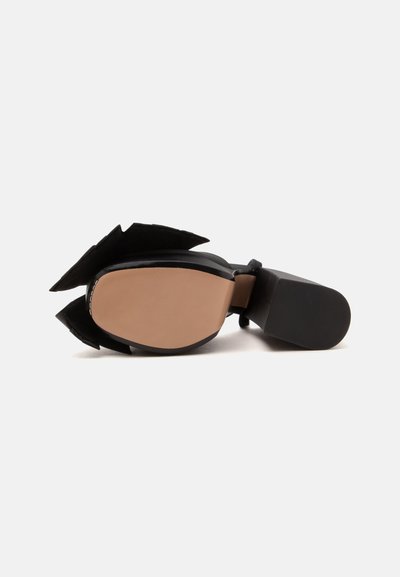 Chaussure noire à talon pointu posée sur le côté, montrant une semelle lisse couleur beige et un petit talon carré sur fond blanc.