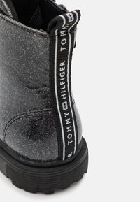 Botín de tobillo negro con purpurina y acabado texturizado, que cuenta con una lengüeta de marca en blanco y negro y una suela de goma gruesa.
