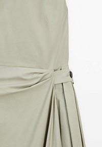 Tissu beige clair avec des plis rassemblés et maintenus par une sangle assortie fixée par un bouton noir.