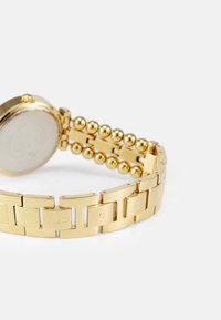 Gold analog watch with a link bracelet featuring flat and beaded segments. The watch has a round case and is engraved with brand letters.