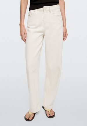 Person iført højtaljede, lige ben off-white jeans parret med sorte og beige thongsandaler, stående mod en ensfarvet baggrund.