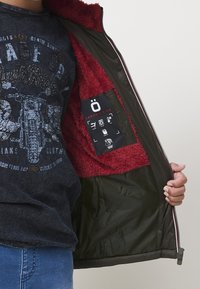 Niño que lleva una camiseta gráfica azul marino y jeans azules, abre una chaqueta verde oscuro forrada con forro de lana roja, mostrando una etiqueta negra de Koroshi Kids en el interior.