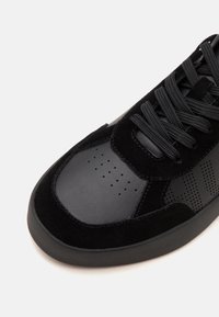 Baskets noires avec dessus en suède texturé et cuir, bout rond perforé et lacets noirs épais sur fond blanc.