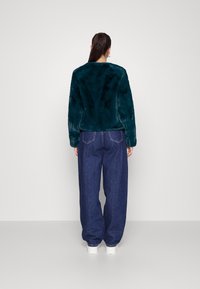 Veste en fausse fourrure turquoise avec un col rond, des manches longues et une texture douce, associée à un jean en denim bleu oversize.