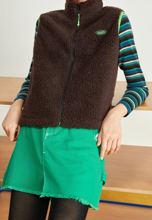 Personne portant un gilet en polaire marron sur des manches longues rayées bleu et vert, une jupe verte à ourlet effiloché et des collants noirs assise sur un banc en bois.