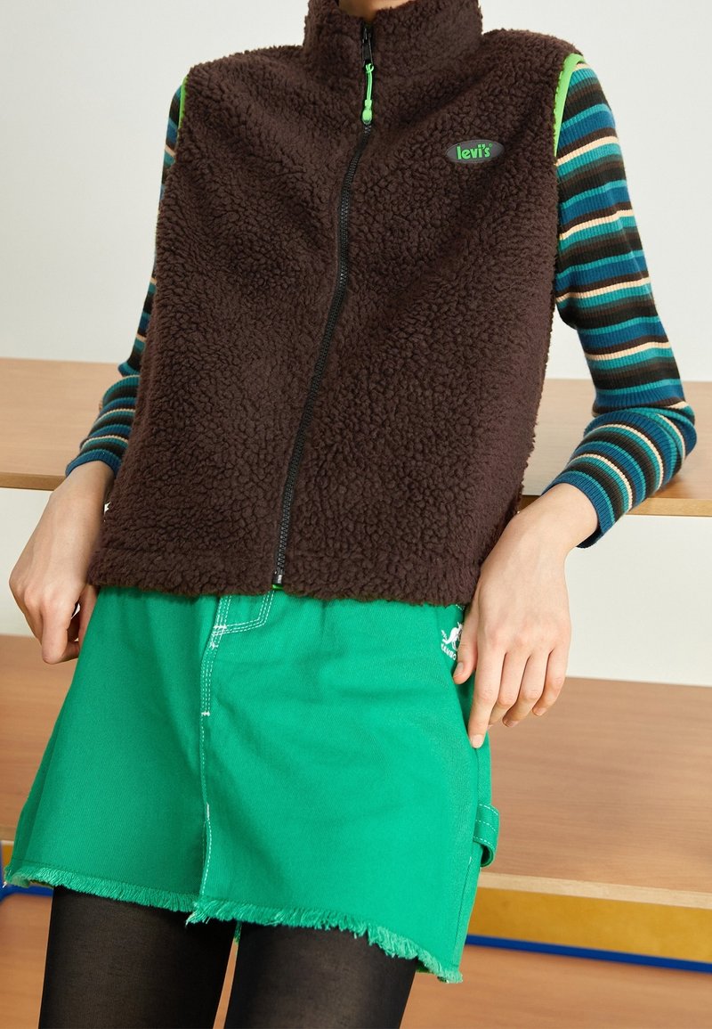 Personne portant un gilet en polaire marron sur des manches longues rayées bleu et vert, une jupe verte à ourlet effiloché et des collants noirs assise sur un banc en bois.
