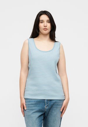 Vrouw met lang donker haar, gekleed in een mouwloos lichtblauw-wit gestreept topje en blauwe spijkerbroek, staand voor een egale achtergrond.